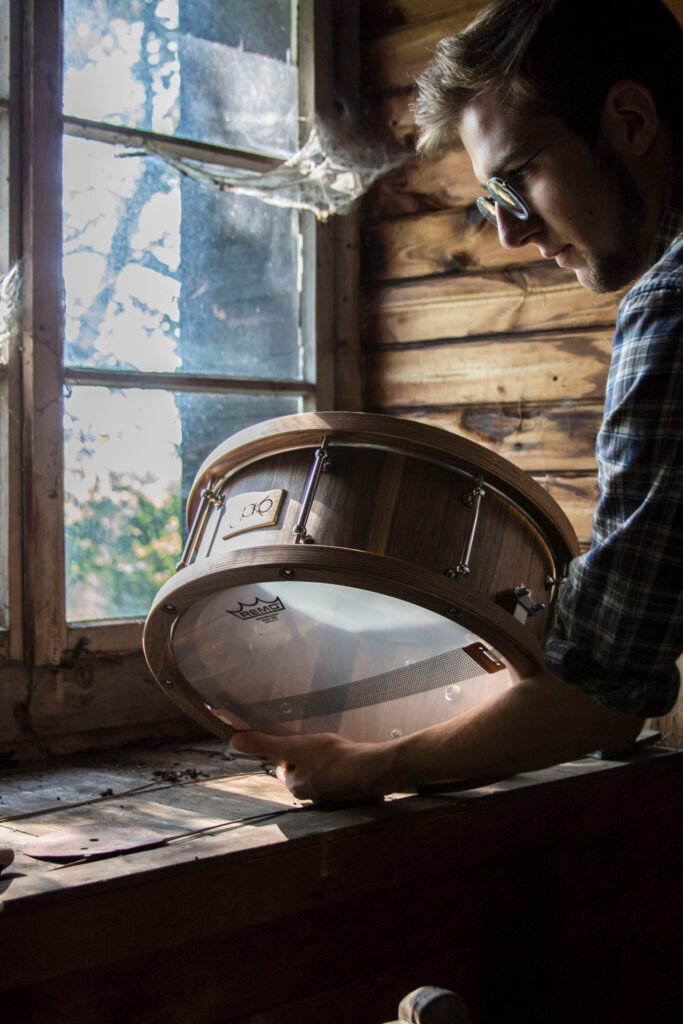 Jonny Kölbl mit einer JoKo Drum am Fenster einer Holzhütte