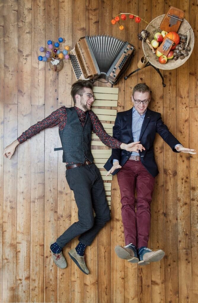 Jonny Kölbl und Leonhard Waltersdorfer liegen mit Harmonika, Marimba-Stäben und Mbira auf einem Holzboden