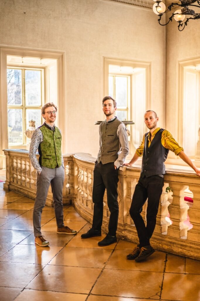 Quetsch ’n’ Vibes in Trachtenwesten lehnen an einer Balustrade in einem hellen Stiegenhaus