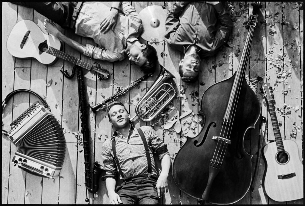 Sturm & Klang liegen in Schwarzweiß mit Harmonika, Kontrabass, Gitarre und Blasinstrumenten auf einem Holzboden