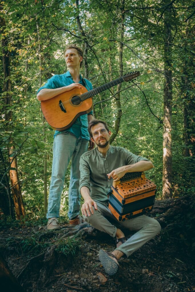 Timmy Gasslander steht mit Gitarre, Jonny Kölbl sitzt mit Harmonika vor einem Baum im Wald