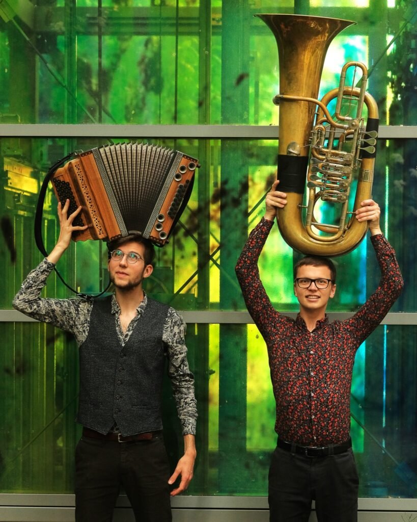 Jonny Kölbl hält eine Steirische Harmonika, Tobias Weiß eine Tuba über den Kopf, vor einer grünen Glasfassade